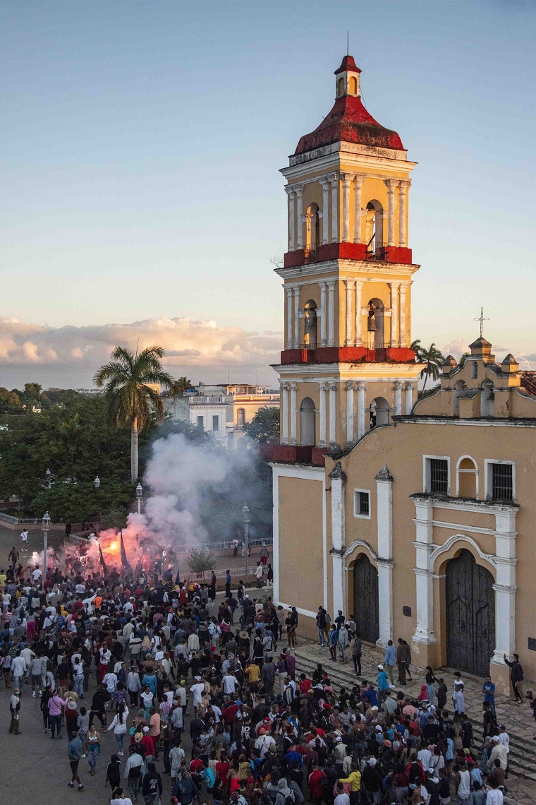 Multitud reunida al atardecer frente a la torre de la iglesia de Remedios entre humo de fuegos artificiales