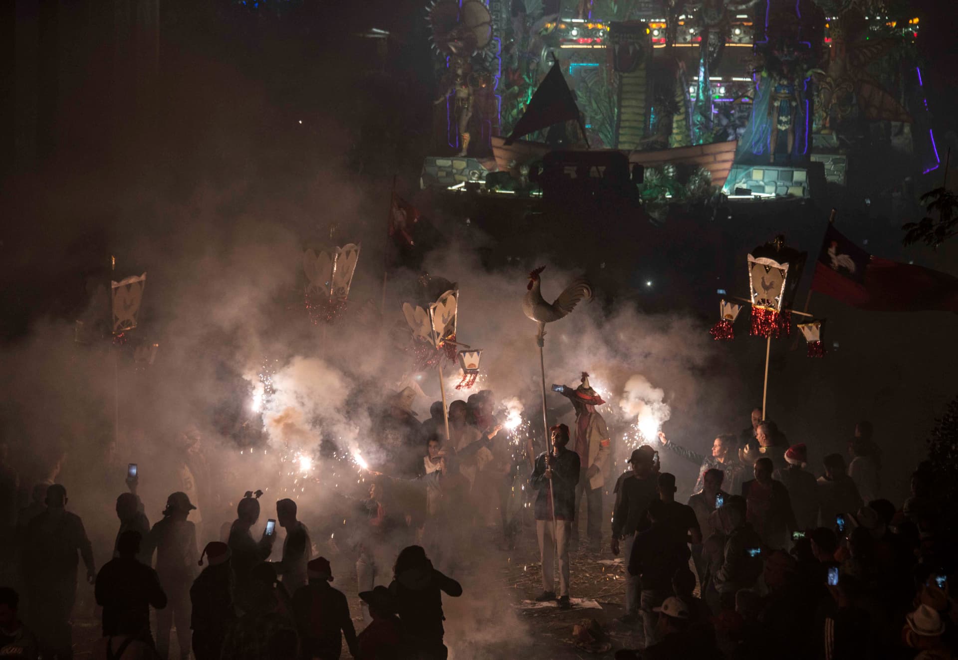 Multitud con banderas y faroles entre el humo de los fuegos artificiales frente a una carroza iluminada